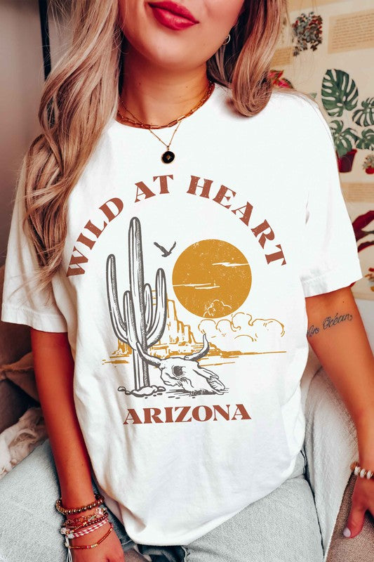 Woman wearing the WILD AT HEART ARIZONA Graphic Tee in white, which features the text "Wild at Heart Arizona" along with an illustration of a cactus, a sun, and a lounging skeleton. This tee is made from 100% cotton and offers unisex sizing for ultimate comfort and style.