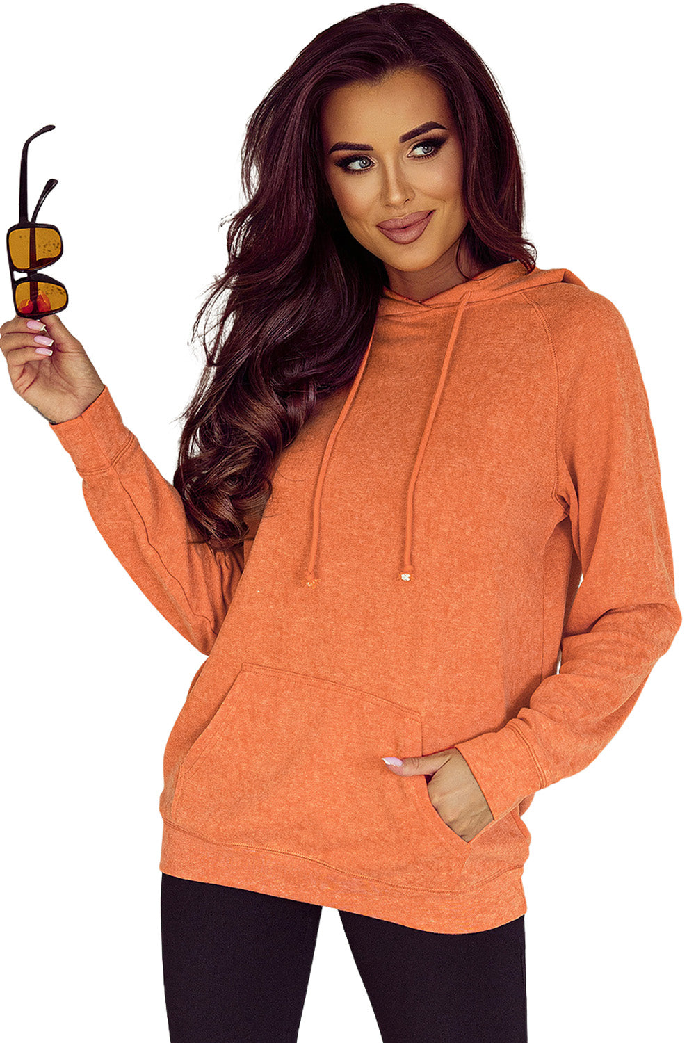 A person wearing an Orange Mineral Wash Kangaroo Pocket Drawstring Pullover Hoodie and sunglasses smiles while looking over their shoulder against a speckled wall backdrop.