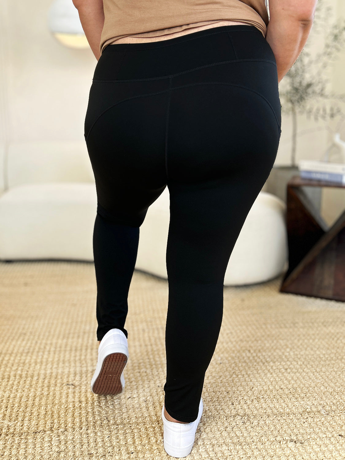 Person wearing Wide Waistband Sports Leggings made of stretchy nylon spandex and white slip-on shoes, standing on a beige carpet with a white couch in the background.