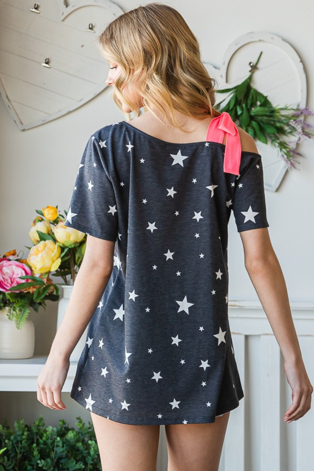 A person wearing the Heimish Full Size Star Print Asymmetrical Neck Short Sleeve Top and denim shorts stands indoors, complemented by floral decor and a heart-shaped display in the background.