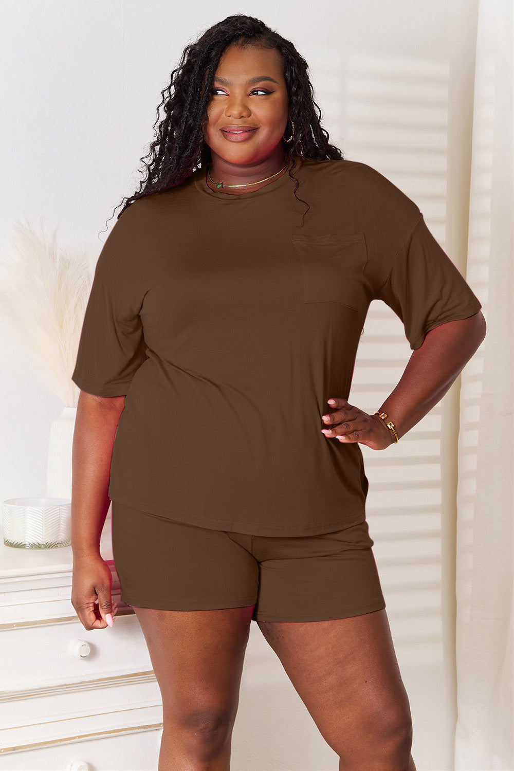 A woman is standing in a brightly lit room, wearing the Basic Bae Full Size Soft Rayon Half Sleeve Top and Shorts Set in a relaxed, stretchy turquoise fabric. She has one hand placed in her pocket and is smiling while looking slightly to the side.