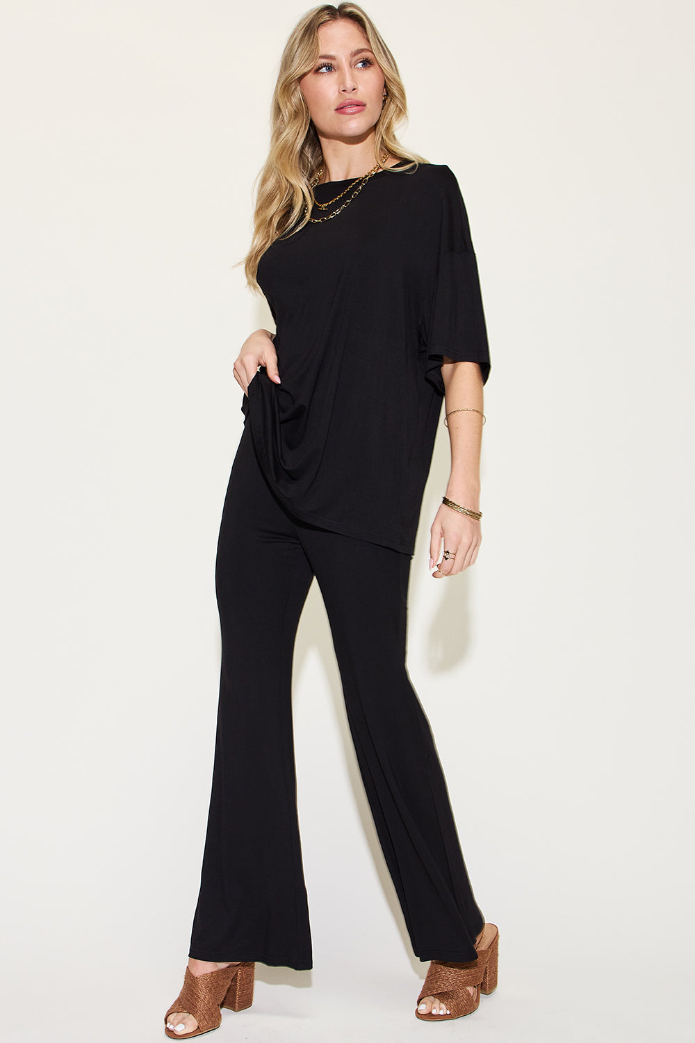 A woman with long, blonde hair wears the Basic Bae Full Size Bamboo Drop Shoulder T-Shirt and Flare Pants Set in black, paired with brown heeled sandals and layered necklaces. She stands against a plain, light background.