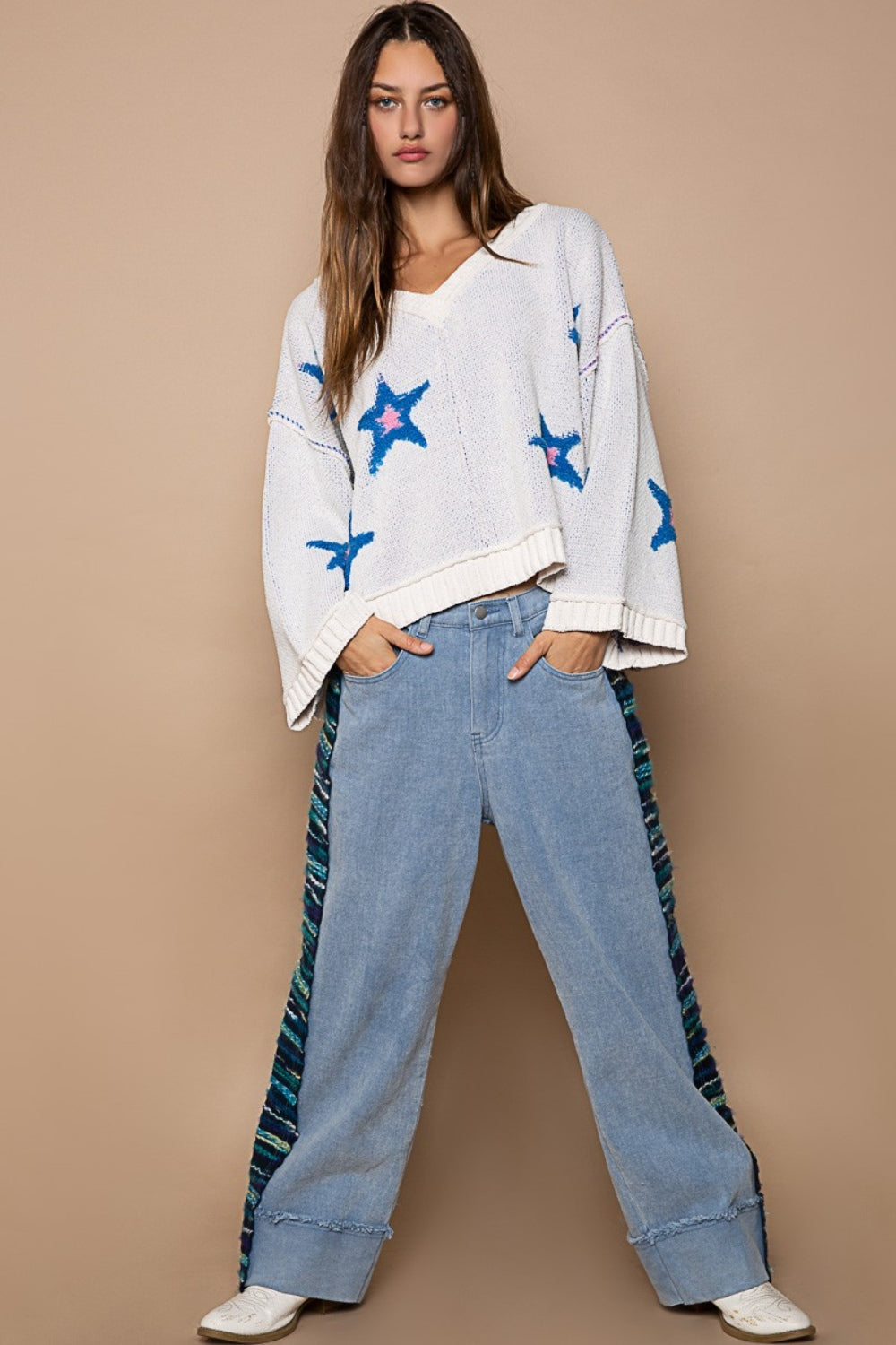 Woman in a POL Long Sleeve Star Patch Sweater with blue color-blocked star berber patches and light blue jeans standing against a beige background.