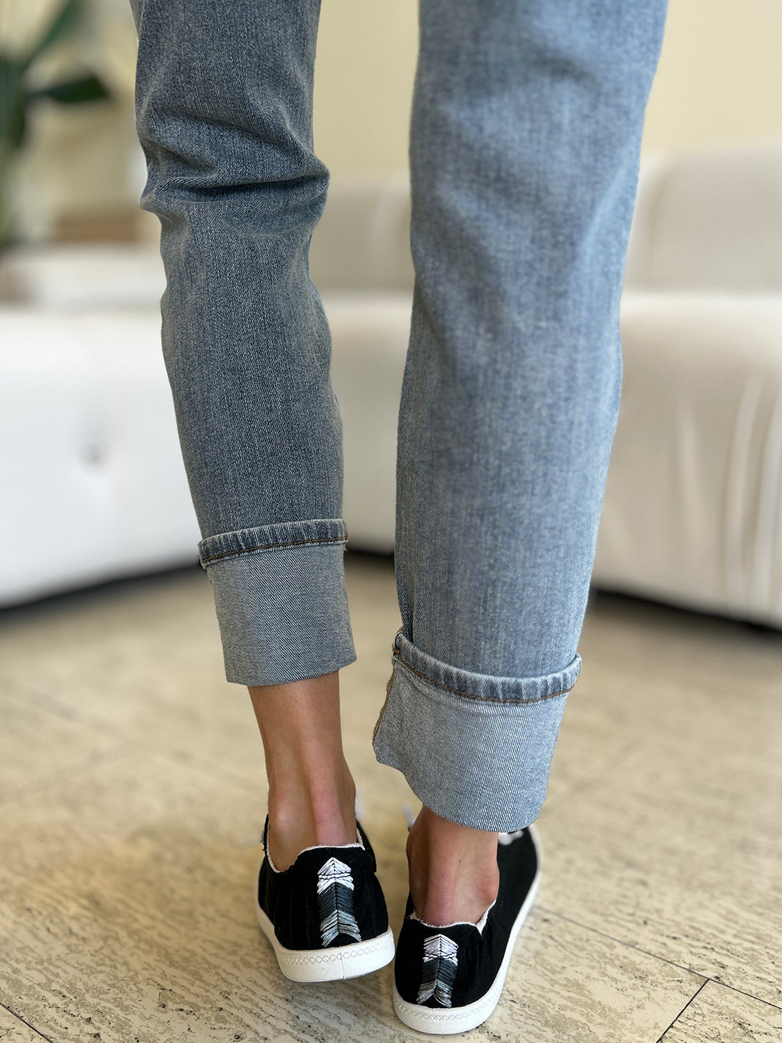 A person wearing Judy Blue Full Size High Waist Cuff Hem Jeans and white slip-on shoes stands in a living room with a white couch and plants in the background.