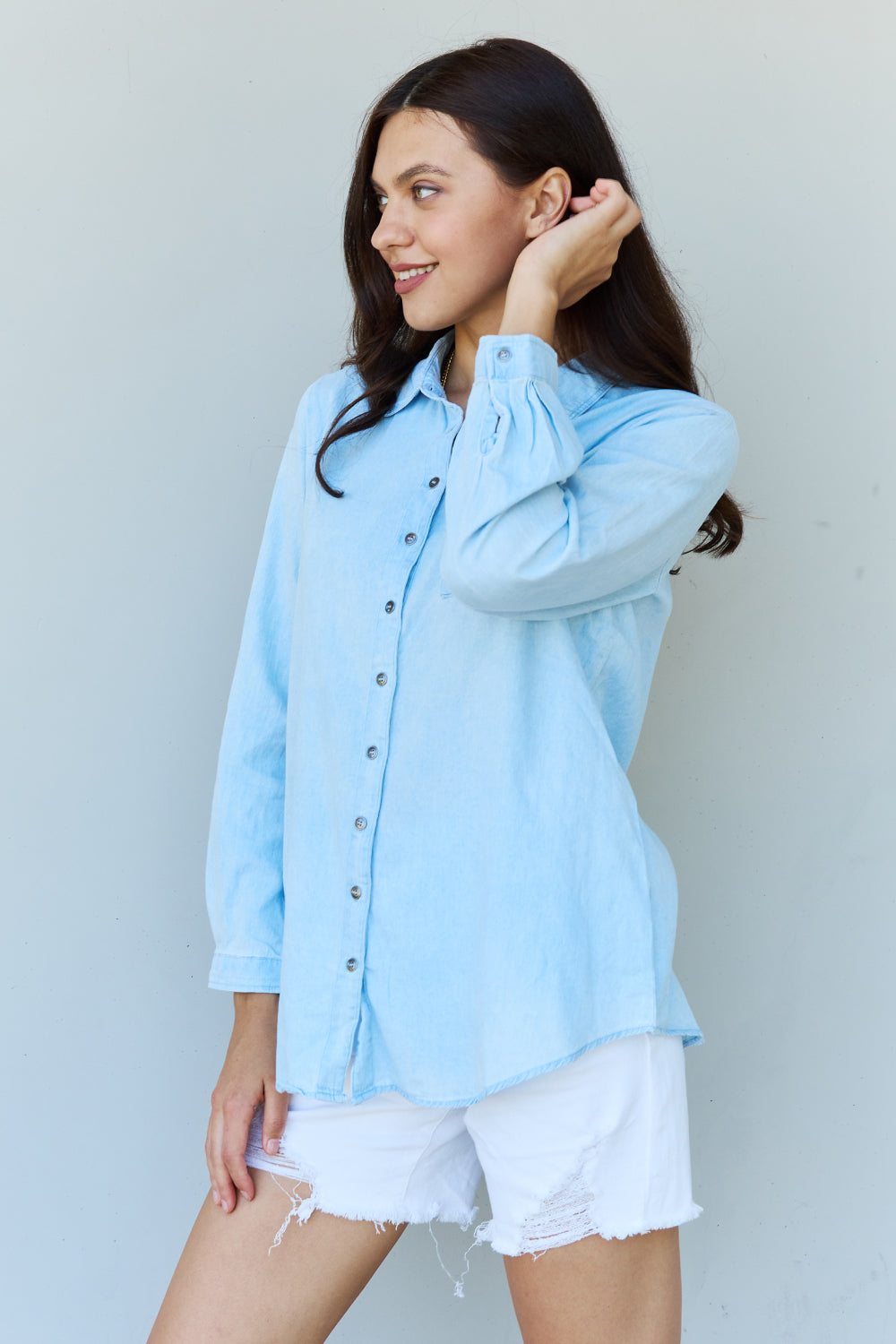 A woman wearing the Doublju Blue Jean Baby Denim Button Down Shirt Top in Light Blue, paired with white ripped shorts, strikes a pose with one hand in her pocket against a plain background, highlighting the classic button-up silhouette.