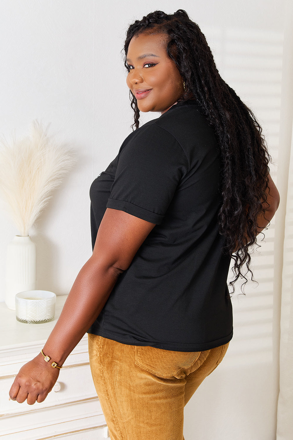 A woman with long dark hair smiles while wearing a trendy black Simply Love Letter Graphic Round Neck T-Shirt featuring the text "ME? Sarcastic? NEVER" and brown corduroy pants. She stands in a room with white walls and decor.