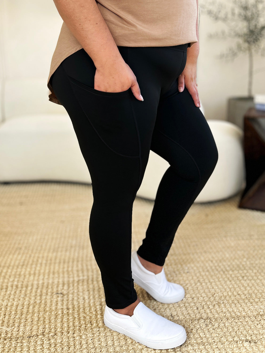 Person wearing Wide Waistband Sports Leggings made of stretchy nylon spandex and white slip-on shoes, standing on a beige carpet with a white couch in the background.