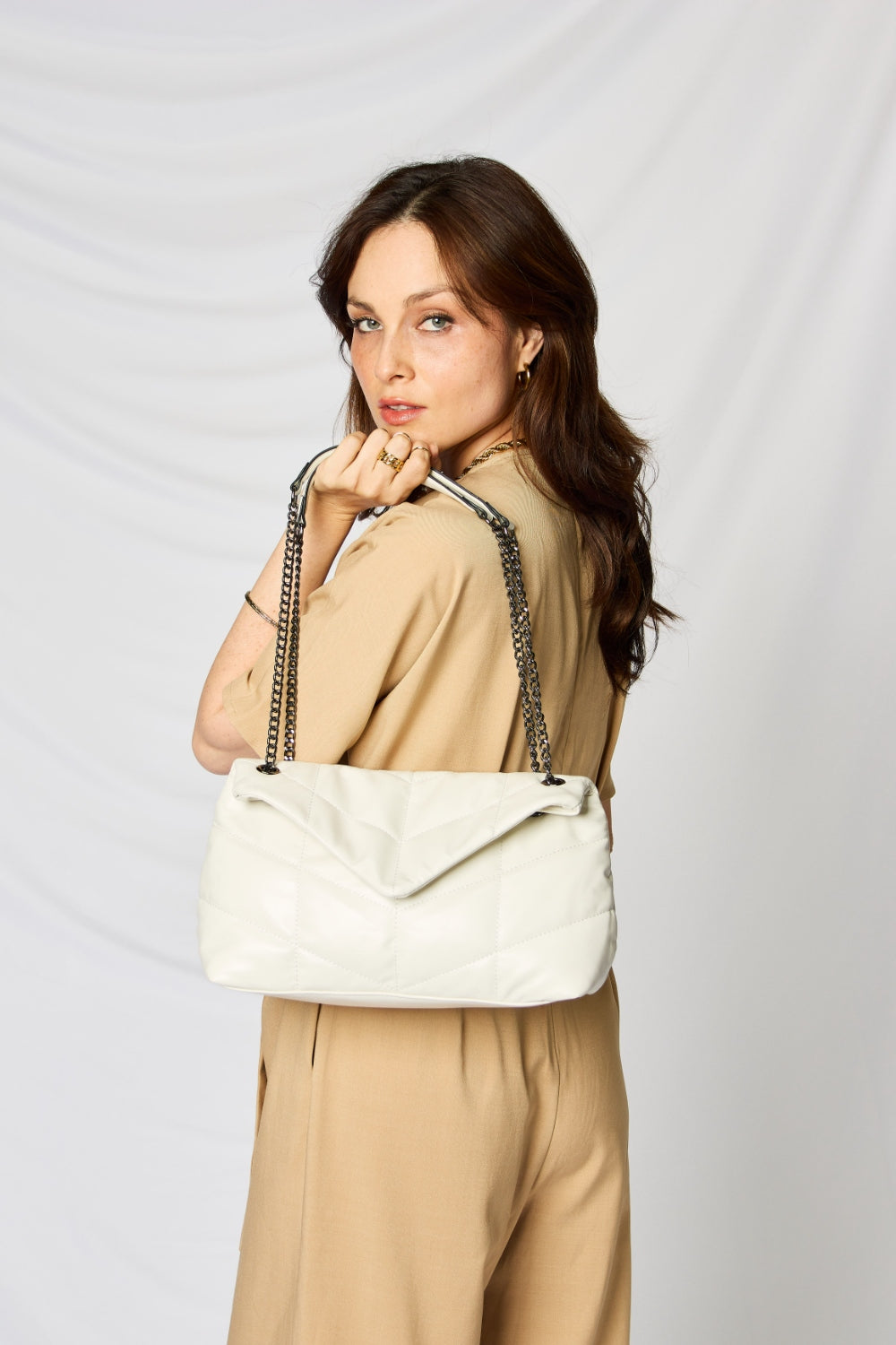 A person holding a stylish SHOMICO PU Leather Chain Handbag over their shoulder against a white background. They are dressed in a beige outfit and looking at the camera, embodying sustainable fashion.