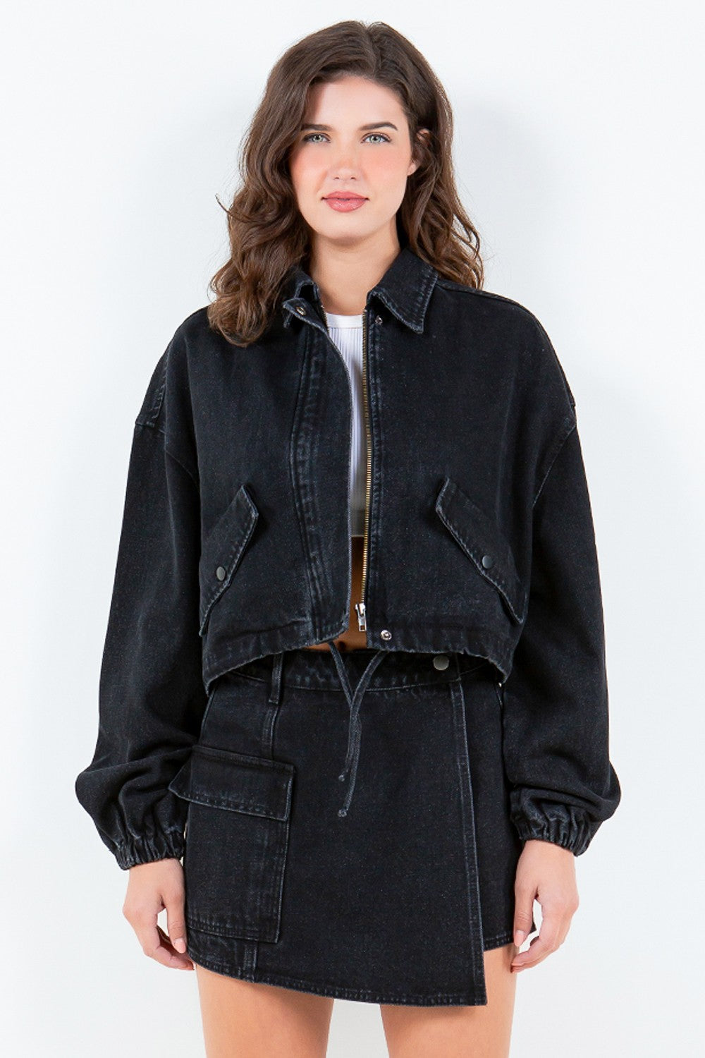 A person with wavy hair is wearing an American Bazi Drawstring Waist Crop Denim Jacket in black, featuring a zippered front closure, paired with a matching black denim skirt.