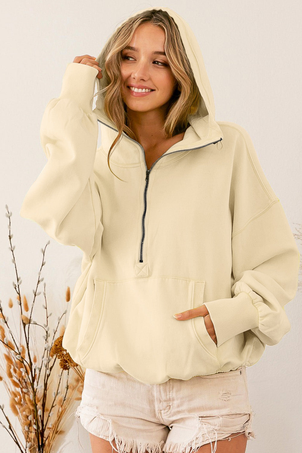 A person wearing a loose fit Beige Ribbed Trim Kangaroo Pocket Zipped Hoodie and frayed shorts stands sideways, with dried plants and a vase in the background.