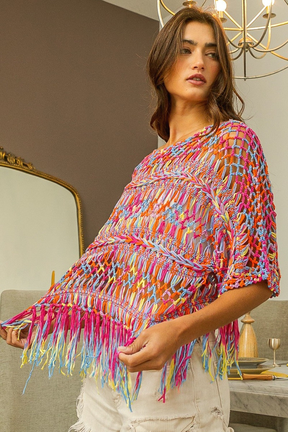 A woman stands indoors wearing the BiBi Openwork Fringed Knit Cover Up over a white outfit, radiating bohemian flair. This versatile piece adds vibrancy to her summer wardrobe.