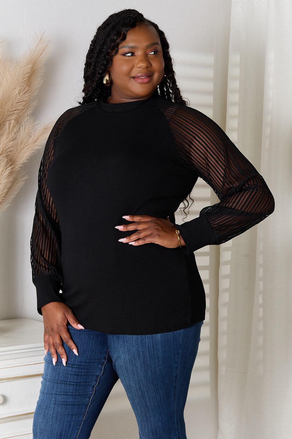 A woman with long dark hair stands indoors wearing a versatile wardrobe piece: the Double Take Round Neck Raglan Sleeve Blouse paired with blue jeans, her right hand in her pocket. A white vase with pampas grass is on the table next to her.