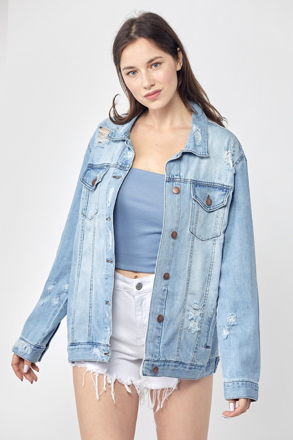 Dressed in the RISEN Full Size Distressed Long Sleeve Denim Jacket, a perfect wardrobe staple, this person effortlessly poses against a plain background while pairing it with a blue top and white shorts.