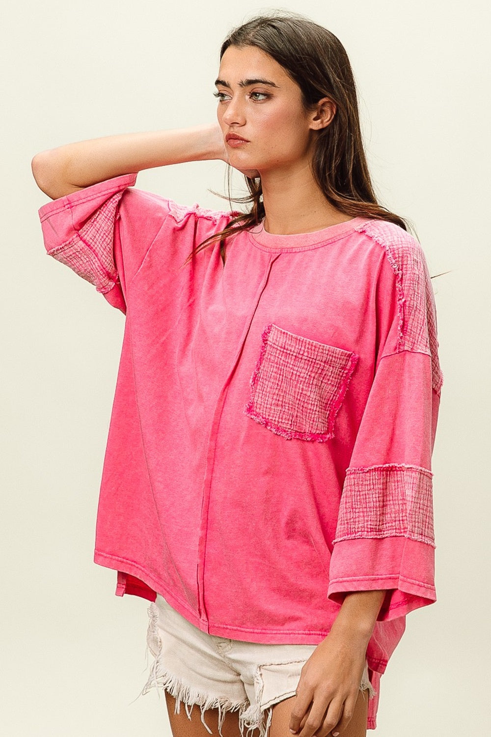 A young woman with long dark hair wears a bright pink BiBi High-Low Washed T-Shirt featuring a front pocket, paired with white frayed denim shorts. She poses against a plain background, effortlessly combining comfort and style in her trendy wardrobe.