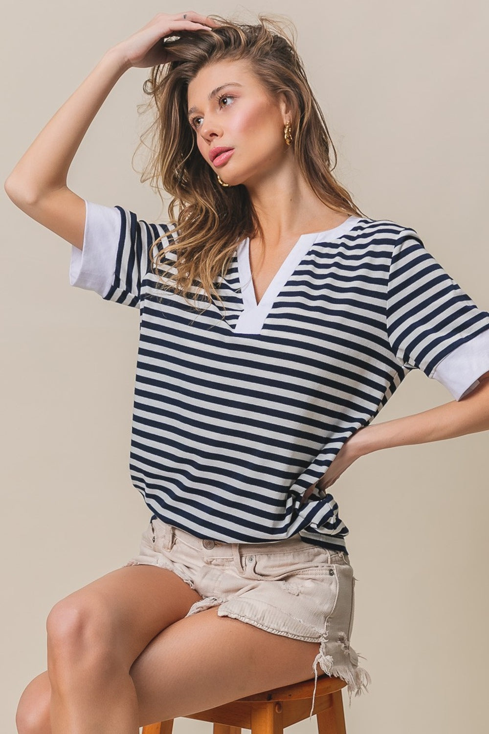 A person with long hair, wearing a BiBi Contrast Striped Notched Knit Top in navy and white and beige shorts, is posing with one hand in their hair, smiling against a beige background.