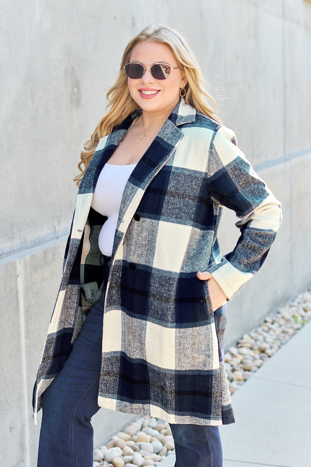 A woman stands on a sidewalk wearing the Double Take Full Size Plaid Button Up Lapel Collar Coat in pink and white over a white top and blue jeans, with her right hand lightly touching her hair. The polyester fabric of the coat makes it durable and perfect for an easy machine wash cold.