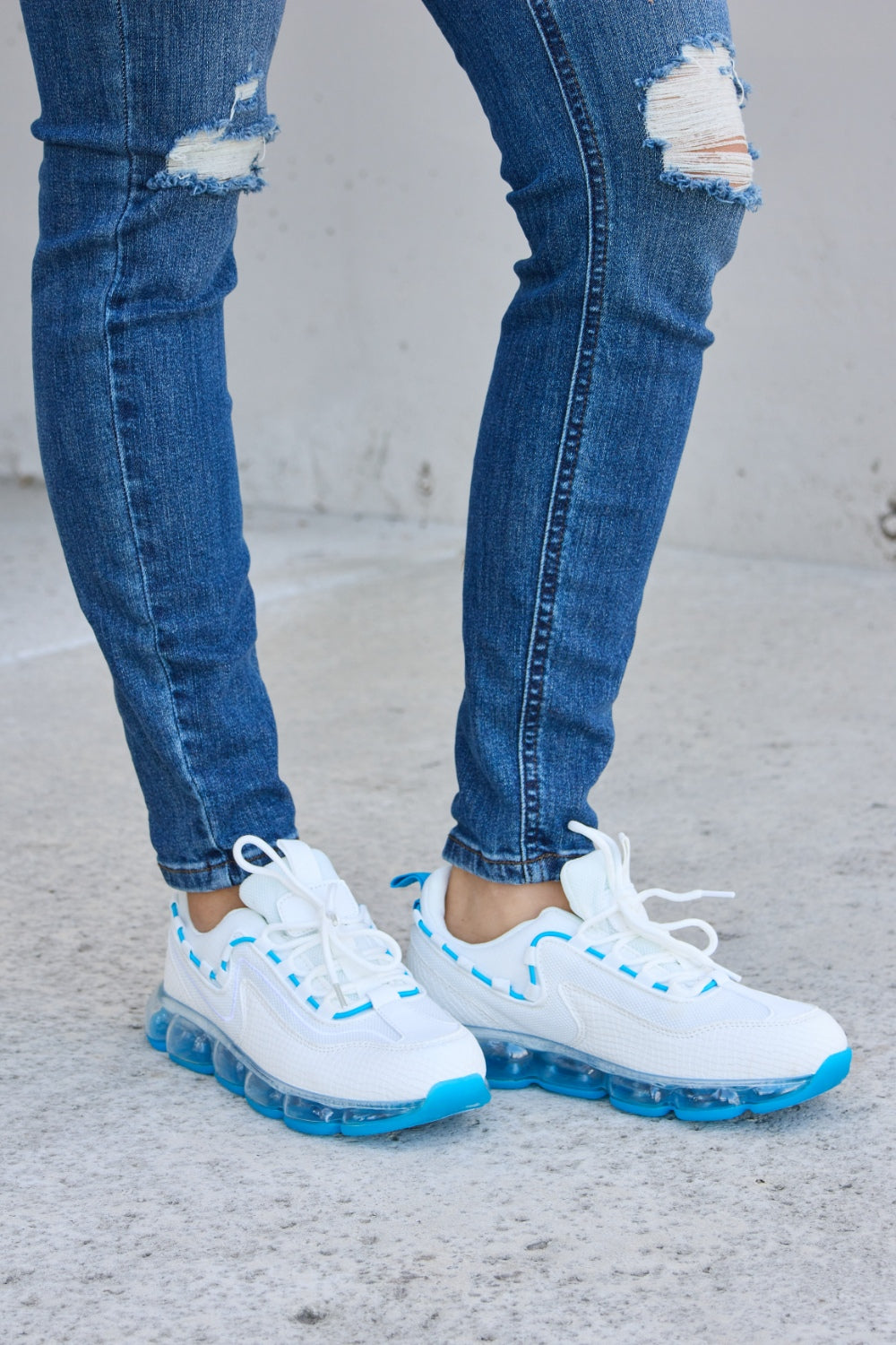 A person wearing ripped jeans and stylish Forever Link Lace-Up Air-Cushioned Athletic Shoes in white with blue accents stands confidently on a concrete surface.