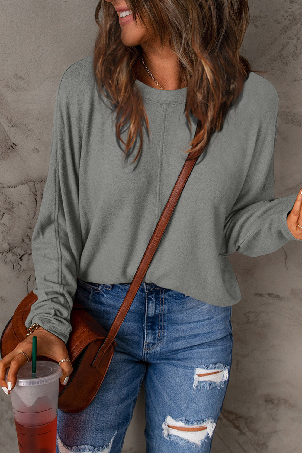 A person wearing a Double Take Full Size Round Neck Long Sleeve T-Shirt in green, blue distressed jeans with a slightly stretchy fit, and a crossbody bag, holding a beverage in a plastic cup.