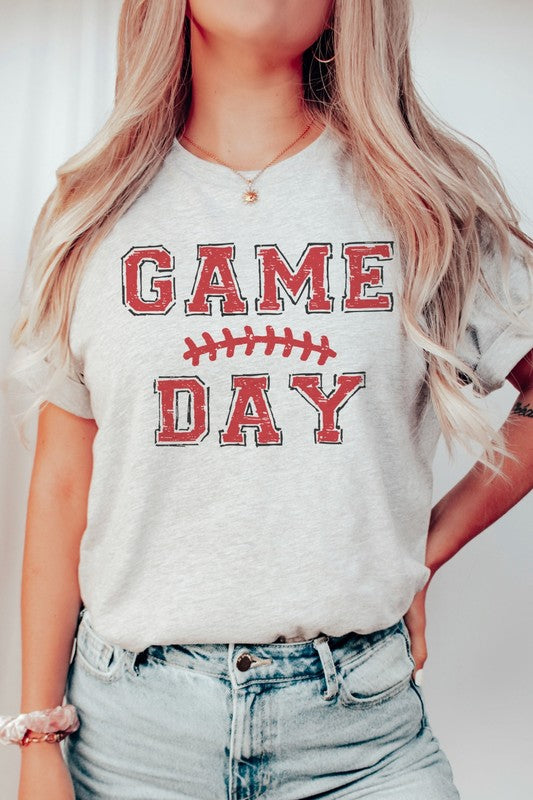 A young woman with long brown hair is wearing a 100% cotton GAME DAY Graphic Tee, featuring a football graphic and "Game Day" text, as she stands against a neutral background.