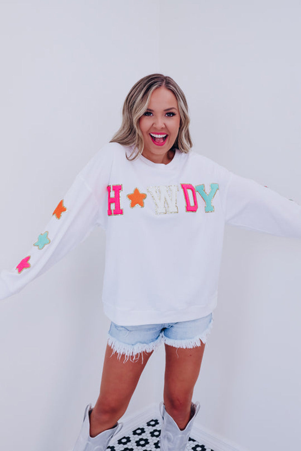 A person poses in the White Howdy Glitter Chenille Patch Graphic Casual Sweatshirt, adorned with colorful star patterns and the letters "WD." She wears light denim shorts and has shoulder-length blonde hair.