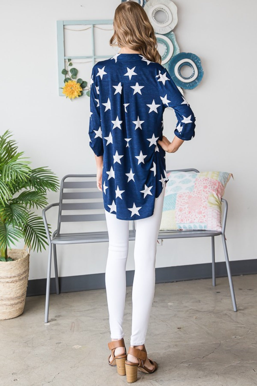 A woman with long hair is sporting the Heimish Full Size Roll-Tab Sleeve Star Print Top in navy blue, paired with white pants, perfect for casual outings. She stands in front of a light-colored wall adorned with round hangings.