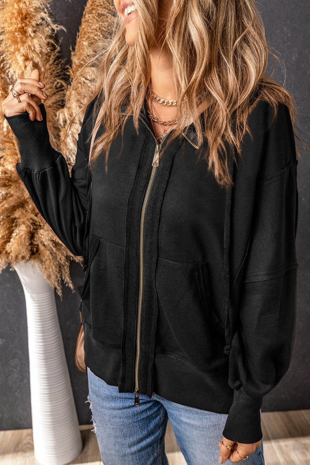 A person with long, wavy blonde hair is shown from the back, wearing a Black Raw Edge Exposed Seam Full Zip Hoodie and blue jeans, and carrying a beige shoulder bag.