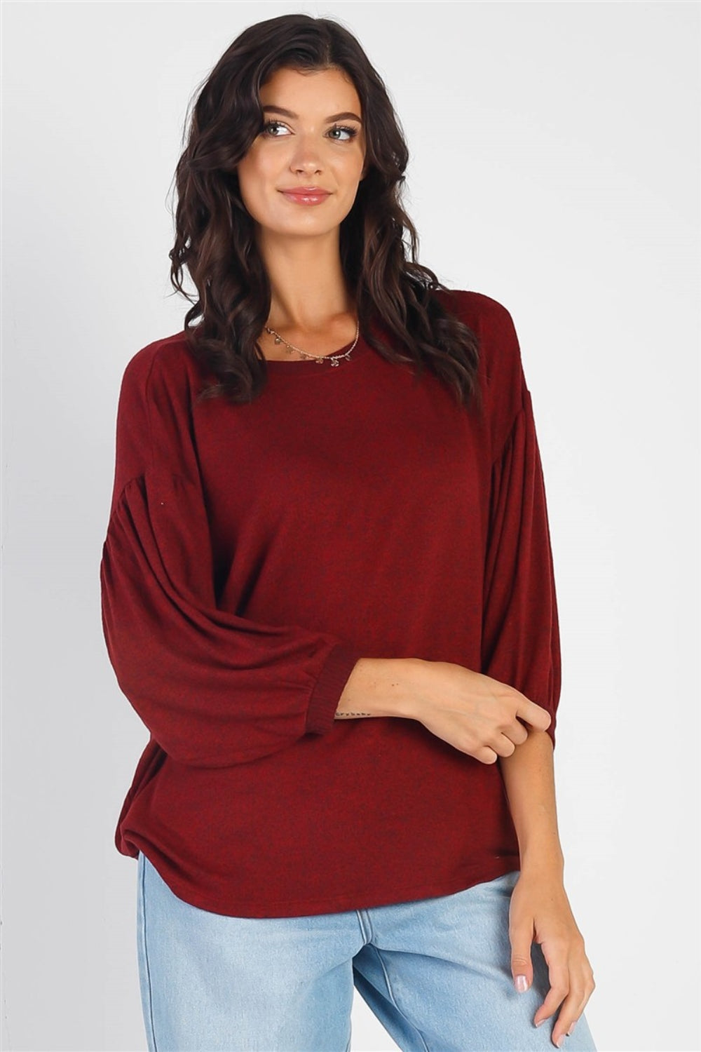 A person with long dark hair stands against a white background, smiling and raising one hand while wearing the trendy and stylish Cherish Apparel Drop Shoulder Puff Sleeve Top in burgundy paired with light blue jeans.