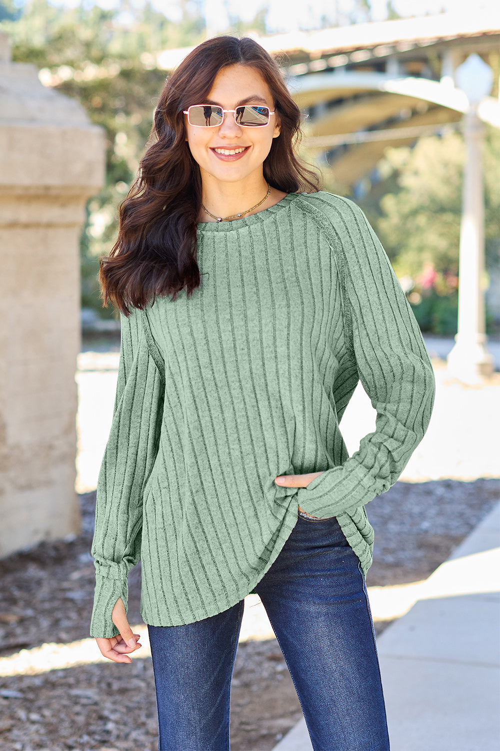 A person with long dark hair and sunglasses stands outdoors, wearing the Basic Bae Full Size Ribbed Round Neck Long Sleeve Knit Top in blue and a pair of jeans. The basic style outfit blends seamlessly with the background of trees and a bridge.