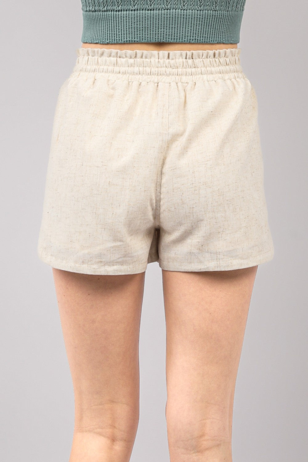 A person wearing VERY J Drawstring Elastic Waist Linen Shorts in beige and a green top. The image focuses on the midsection, with the eco-friendly shorts featuring front pockets and a slight texture.