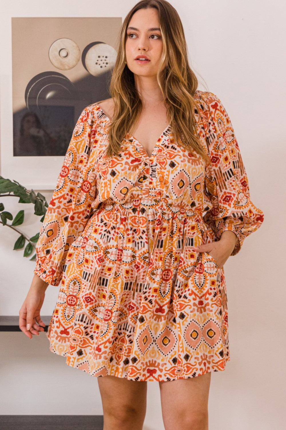 A person stands wearing the ODDI Full Size Printed Ruff Sleeve Romper with Pockets, featuring vibrant orange, red, and yellow designs. They are positioned in front of a wall adorned with framed art and a plant.