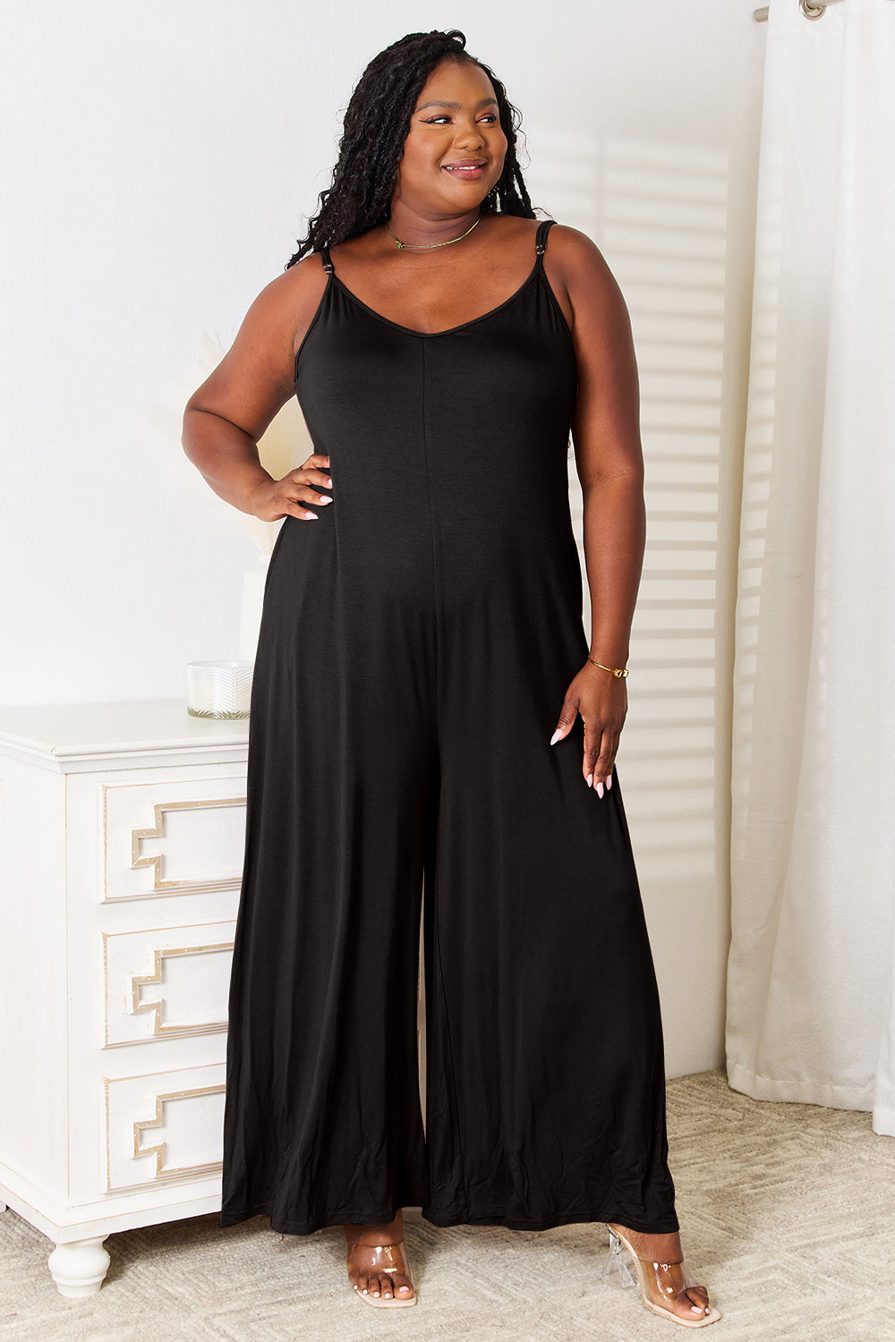 A woman stands indoors wearing a fashionable Double Take Full Size Soft Rayon Spaghetti Strap Tied Wide Leg Jumpsuit. She has one hand resting on a white dresser and is smiling.