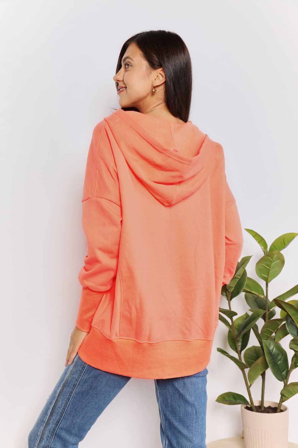 A woman with long hair wears a Double Take Quarter-Snap Dropped Shoulder Hoodie in solid orange. She is standing, with one hand touching her hair and the other resting in the pocket of her jeans, exuding a casual vibe.