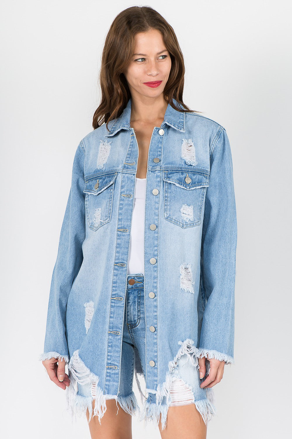 A woman sporting an American Bazi Distressed Frayed Hem Denim Jacket in light blue, paired with a white top and ripped denim shorts, stands against a plain background, exuding a vintage vibe with the frayed hem details.