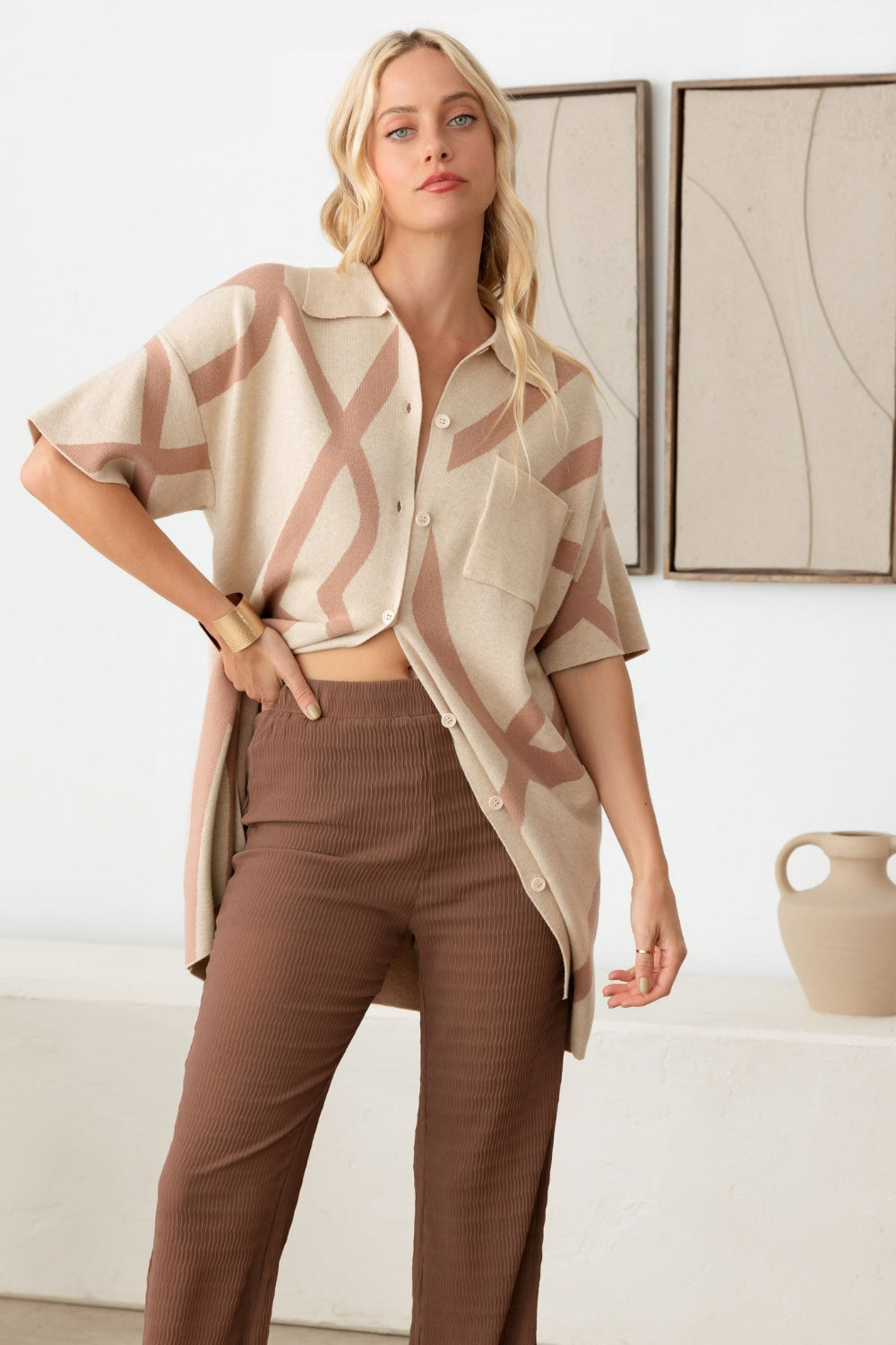 A woman with long blond hair wearing the Le Lis Abstract Collared Button Down Sweater Dress stands in a minimalist room with a wooden chair and ceramic vase—her versatile piece adds an abstract touch to the setting.