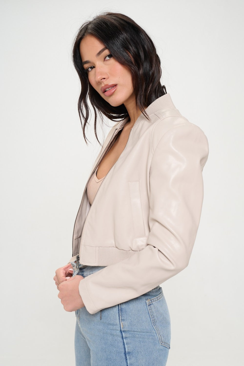 A woman wearing a Coalition LA Zip Up Cropped Bomber Jacket over a ribbed beige top and light blue jeans stands facing the camera, embodying sustainable fashion.