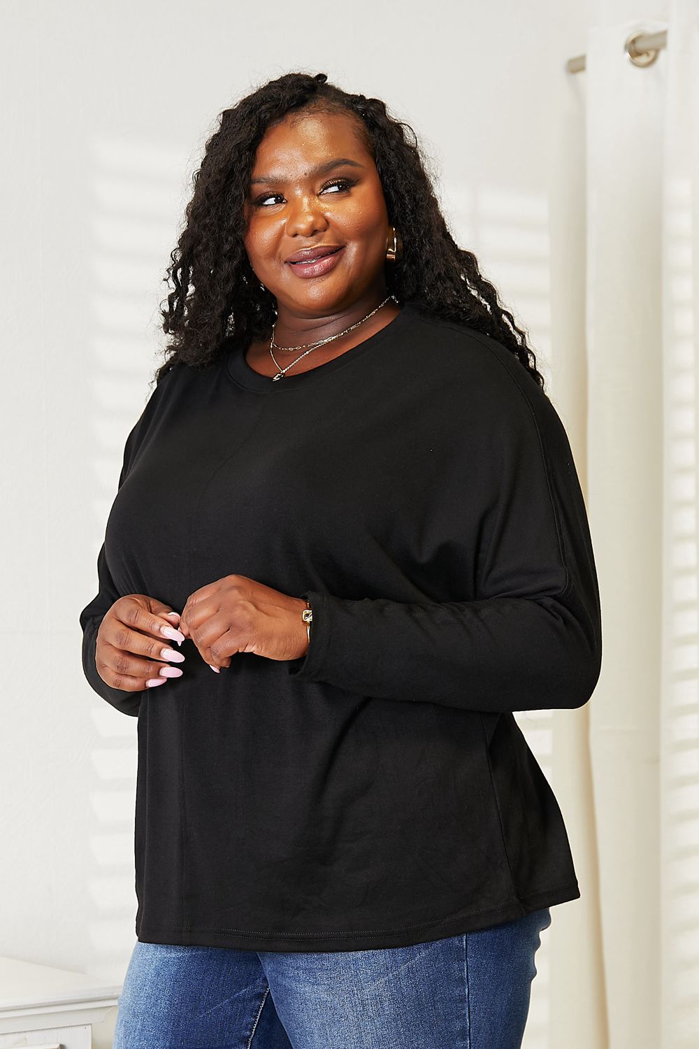 A person with long hair wearing a Double Take Seam Detail Round Neck Long Sleeve Top in solid black and blue jeans stands with one hand in their pocket, looking downward and smiling.