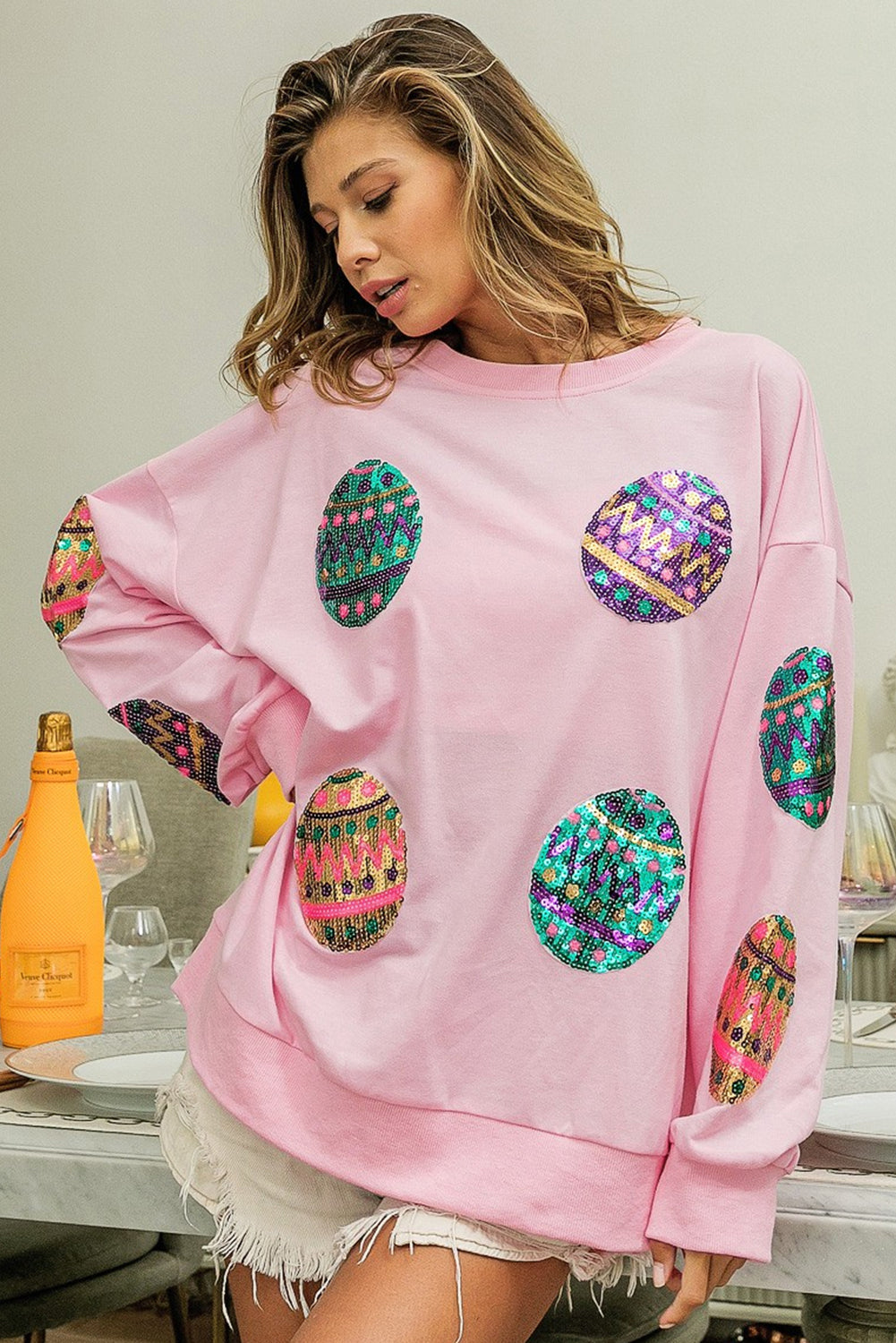 A woman with long, wavy hair wears a Pink Sequined Easter Egg Drop Shoulder Oversized Sweatshirt featuring colorful, patterned circles. She is seated in a dining area with a table set with glasses, plates, and two bottles, one of which is orange.