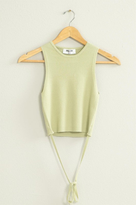 The Good Times Ribbed Sweater Tank Top, featuring a light green color, cropped hem, and back tie, hangs on a wooden hanger against a beige wall.