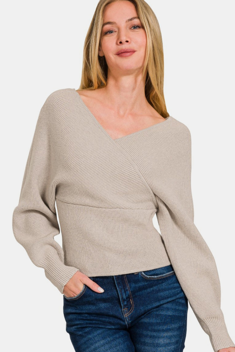 A woman with long blonde hair is smiling with one hand in her pocket against a plain white background, wearing the Zenana Cross Wrap Rib Long Sleeve Sweater in beige and blue jeans—a perfect layering option for any season.