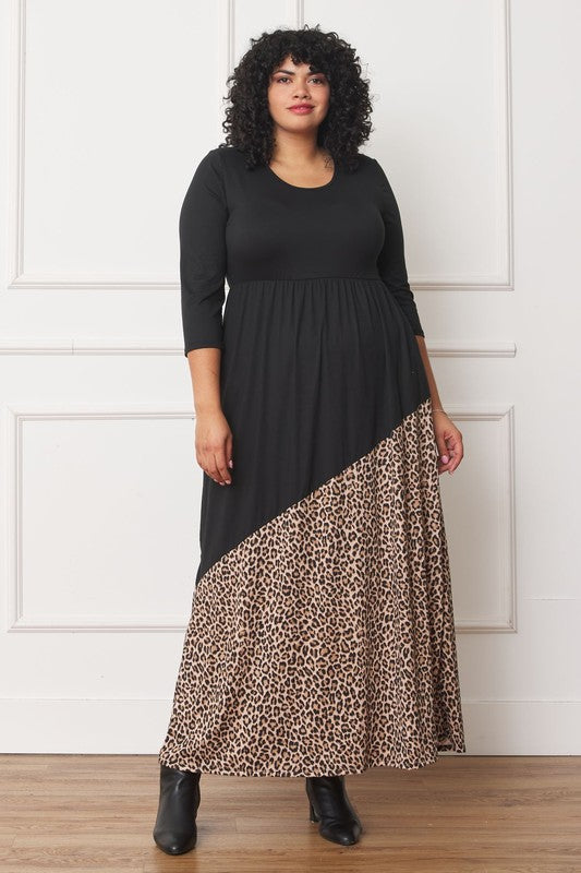 A woman with curly hair is wearing the Asymmetrical Leopard Accent Maxi Dress, featuring a long black design with a floral patterned section at the bottom. She stands against a white paneled wall, her hands tucked in her dress pockets, showcasing its elegant design.