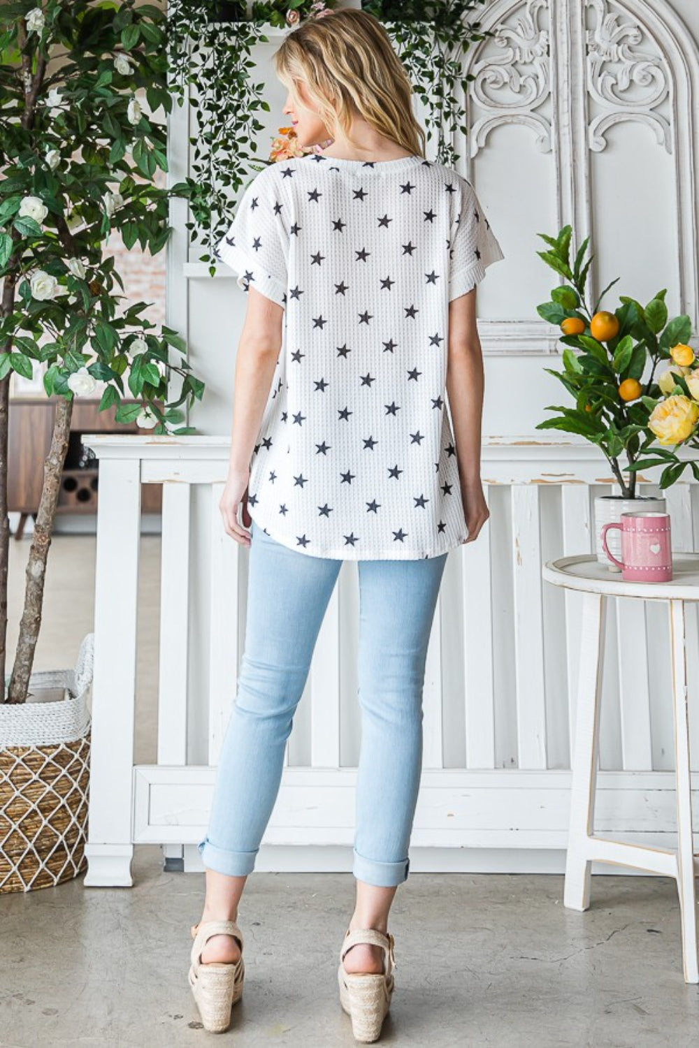 A person with long blonde hair is wearing the Heimish Full Size Star Print Short Sleeve V-Neck Waffle Knit T-Shirt. They are standing indoors near a plant.