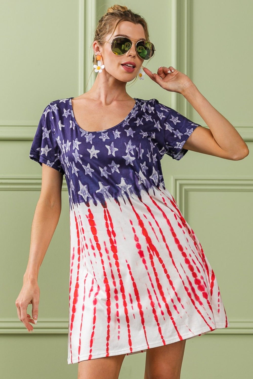 A person wearing the BiBi American Flag Theme Tee Dress, accessorized with sunglasses and earrings, stands against a light green wall with decorative molding.