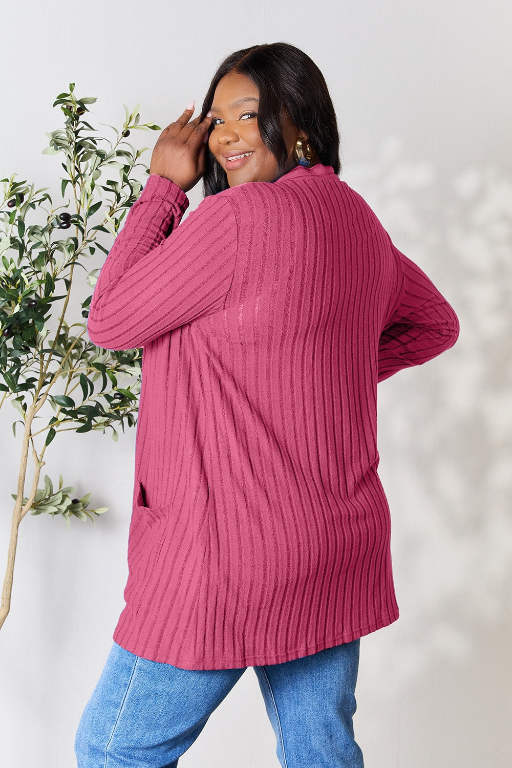 A woman in a Basic Bae Full Size Ribbed Open Front Cardigan with Pockets and jeans, made from stretchy material, stands in front of a plant, smiling slightly and looking to the side.