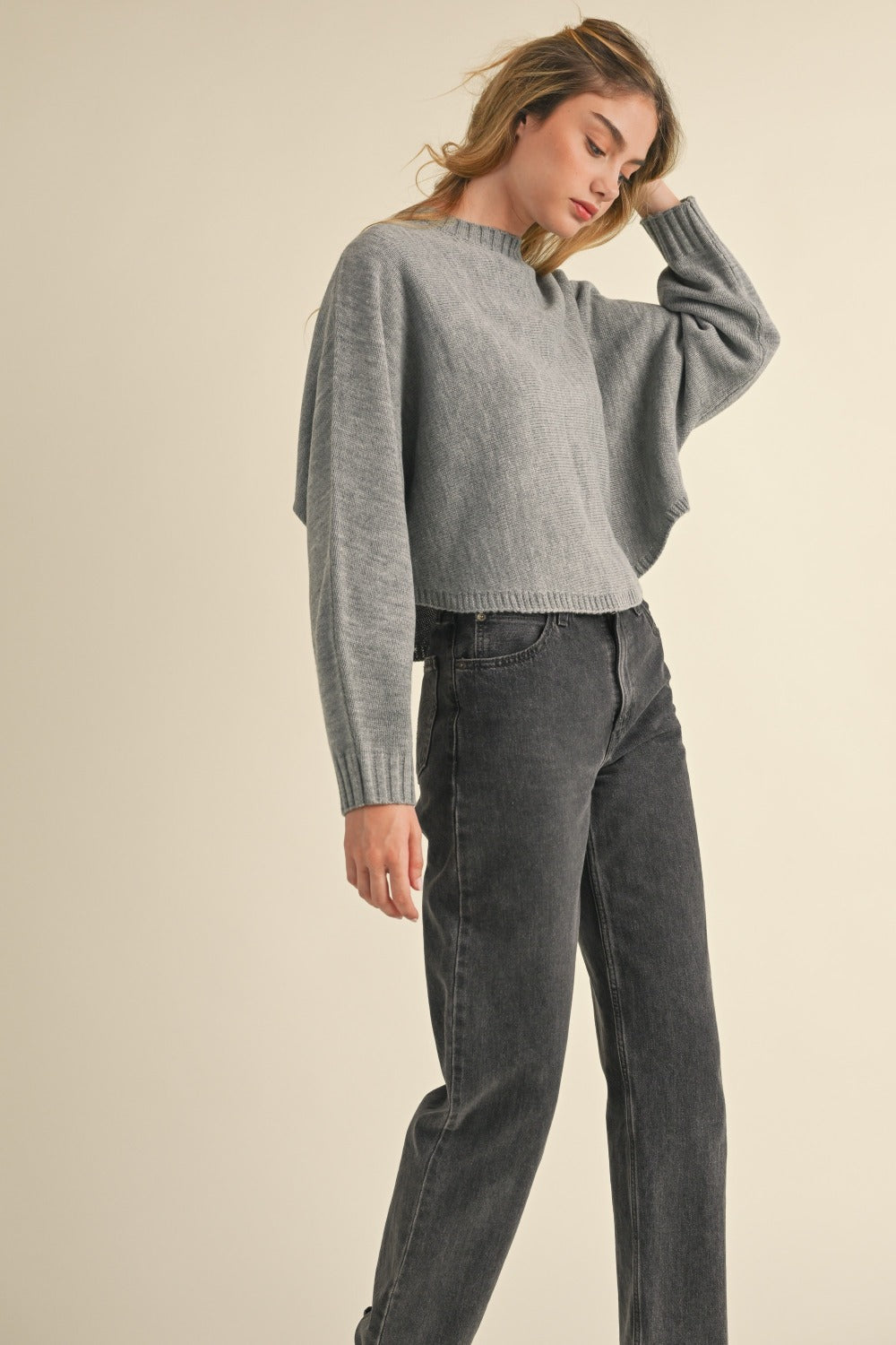 A person with long, light brown hair is facing away, wearing a Mable Round Neck Dolman Sleeve Cropped Sweater in grey paired with dark jeans, against a neutral background.