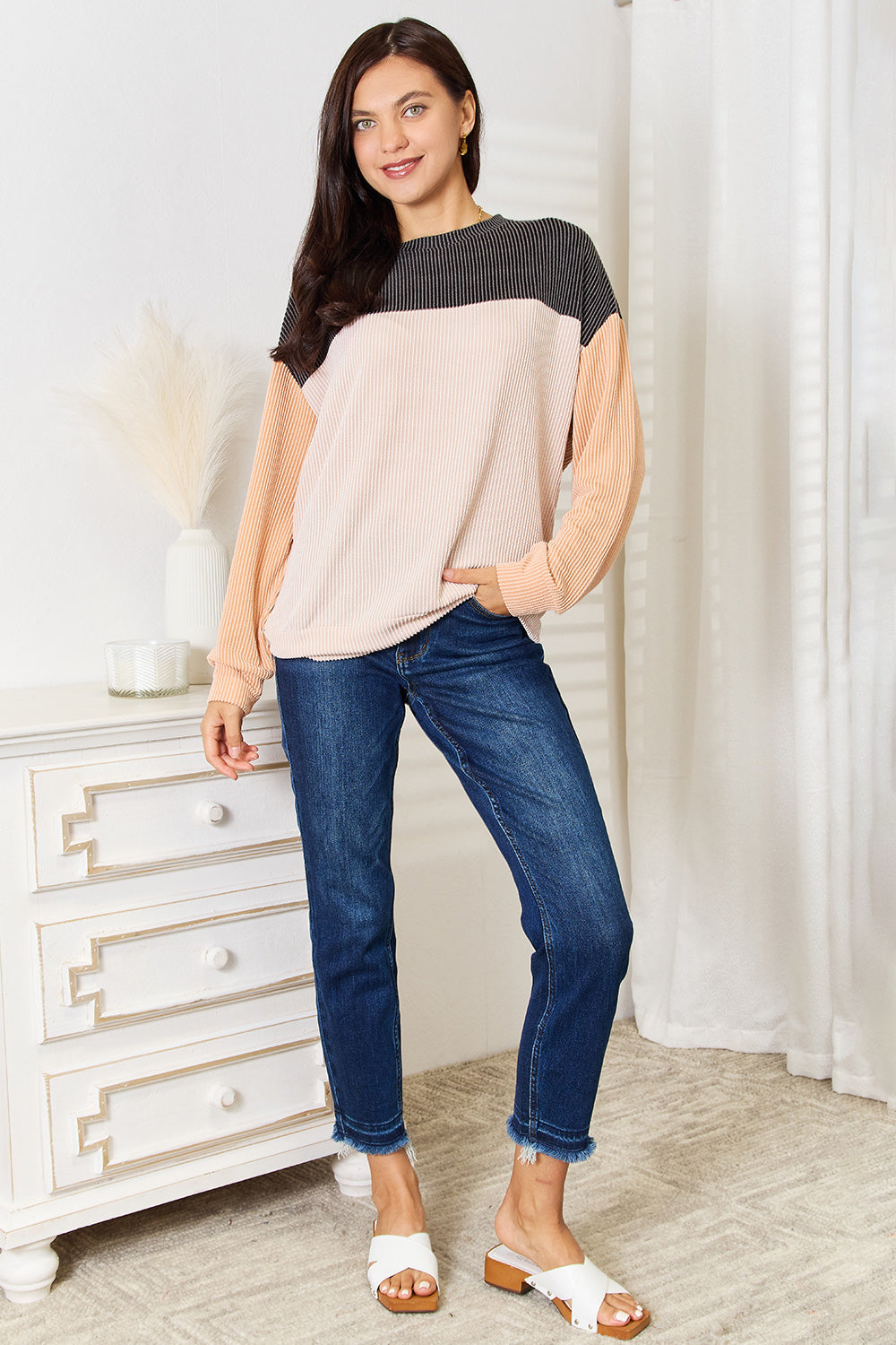 A woman with long brown hair stands indoors, smiling, and wearing a Double Take Color Block Dropped Shoulder T-Shirt paired with jeans. She is positioned near a small table with a white vase containing dried pampas grass, epitomizing the perfect casual wardrobe.