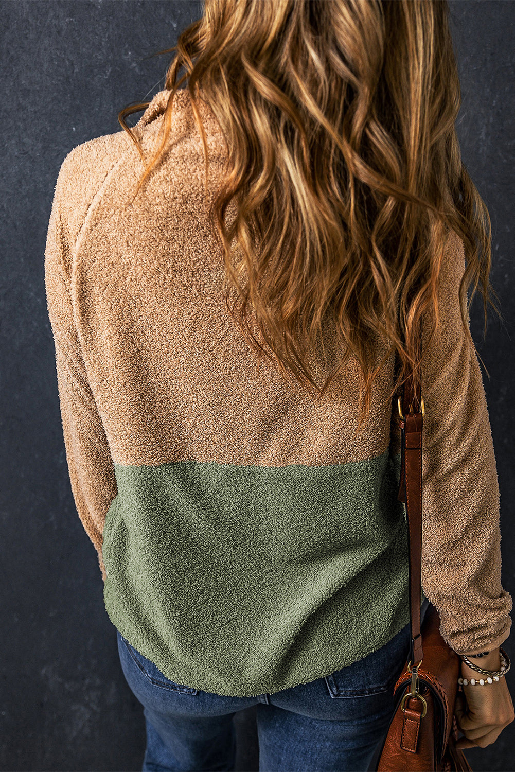 A person with wavy hair, wearing a smoke gray two-tones color block half zip fleece sweatshirt and carrying a brown leather bag is shown from the back against a dark background.