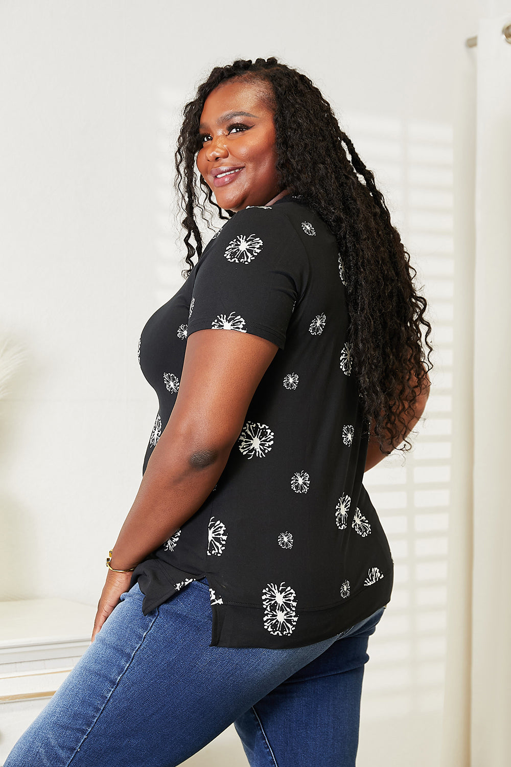 A woman with long braided hair is smiling and wearing the Double Take Dandelion Print Round Neck T-Shirt paired with blue jeans. She stands in front of a light-colored wall, embodying nature-inspired fashion.