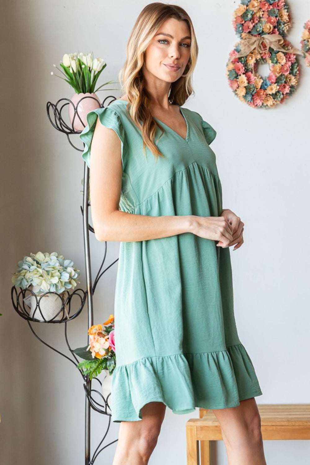 A person in a Heimish Full Size Short Sleeve V Neck Ruffled Hem Dress stands indoors, elegantly holding a hat. The floral decor in the background complements the dress's feminine charm.