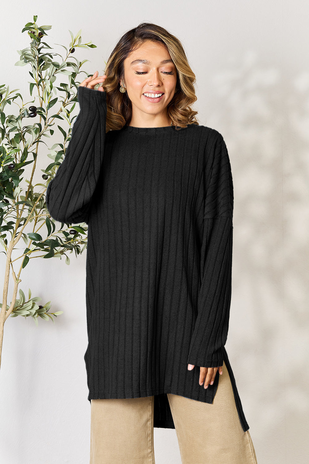 A person with shoulder-length hair smiles while wearing a beige ribbed sweater called the Basic Bae Full Size Ribbed Round Neck Long Sleeve Slit Top, along with matching beige pants, showcasing a basic style. They stand next to a green plant.