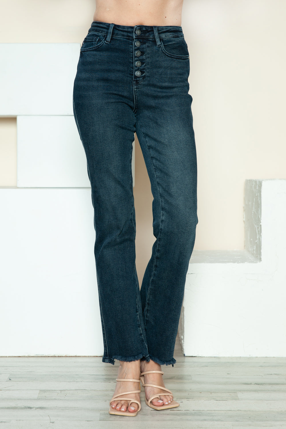 A person is modeling the Judy Blue Full Size Button Fly Hem Destroy Straight Jeans, high-waisted with distressed detailing and frayed hems, paired with light-colored open-toe heels. The background includes white blocks and a light-colored wall.